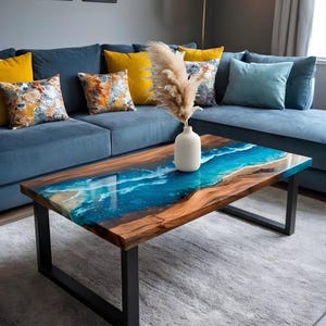 May include: A rectangular coffee table with a wood grain surface and a blue epoxy resin river design. The table has black metal legs and is surrounded by a blue sofa with yellow and patterned pillows. A white vase with dried flowers sits on the table.