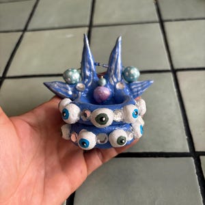 Peut inclure: Un bougeoir en céramique bleu fait à la main avec un design unique. Le bougeoir présente de nombreux petits yeux peints autour de la base et des éléments décoratifs sur le dessus. Le bougeoir est tenu dans une main.