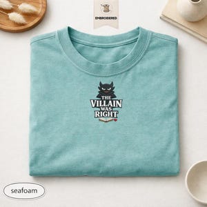 Op de afbeelding: Een zeegroen t-shirt met ronde hals en een geborduurd ontwerp van een zwart duivelsfiguur boven de tekst "THE VILLAIN WAS RIGHT" en een klein icoontje van een open boek met een minuscuul rood hartje. Het ontwerp is gecentreerd op de borst.