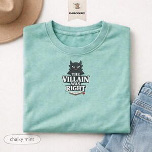Op de afbeelding: Een krijtachtig mintgroen t-shirt met ronde hals en een geborduurd ontwerp van een zwart gehoornd figuur boven de tekst "THE VILLAIN WAS RIGHT" en een icoon van een open boek met een klein rood hartje, gecentreerd op de borst.