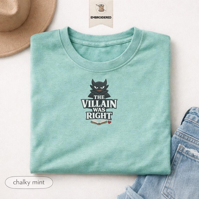 Op de afbeelding: Een krijtachtig mintgroen t-shirt met ronde hals en een geborduurd ontwerp van een zwart gehoornd figuur boven de tekst "THE VILLAIN WAS RIGHT" en een icoon van een open boek met een klein rood hartje, gecentreerd op de borst.