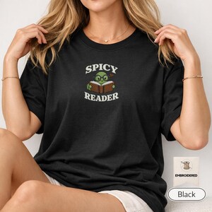 Może przedstawiać: Czarny t-shirt z krótkim rękawem z haftowaną grafiką przedstawiającą zielonego żółwia w okularach czytającego otwartą książkę. Napis "SPICY READER" jest wyhaftowany białą nicią nad i pod ilustracją na wysokości klatki piersiowej.