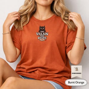 Op de afbeelding: Een T-shirt in gebrand oranje met een centraal geborduurd ontwerp van een zwart duivelsfiguur boven de tekst "THE VILLAIN WAS RIGHT" en een icoon van een open boek met een klein rood hartje. Het T-shirt heeft korte mouwen en een ronde hals.
