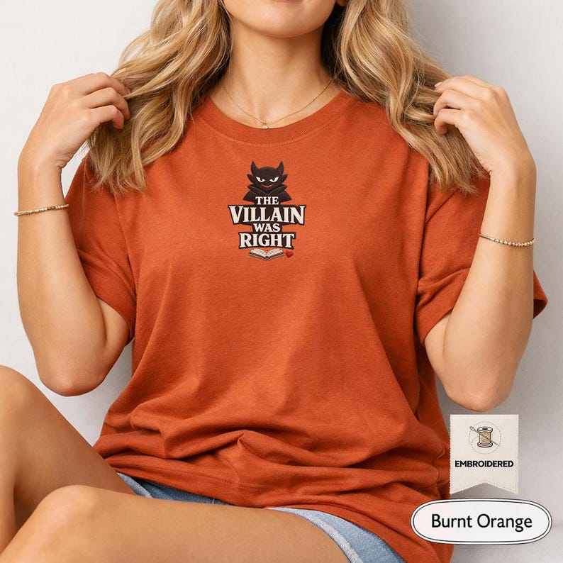 Op de afbeelding: Een T-shirt in gebrand oranje met een centraal geborduurd ontwerp van een zwart duivelsfiguur boven de tekst "THE VILLAIN WAS RIGHT" en een icoon van een open boek met een klein rood hartje. Het T-shirt heeft korte mouwen en een ronde hals.