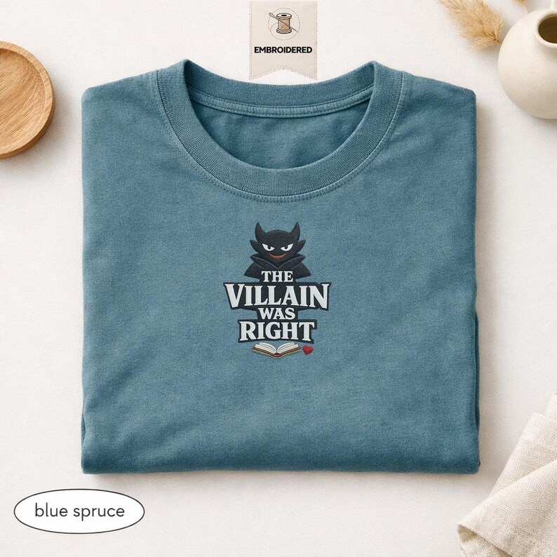 Op de afbeelding: Een blauw-sparrenkleurig t-shirt met een geborduurde afbeelding van een zwart gehoornd personage boven de witte tekst "THE VILLAIN WAS RIGHT" en een icoon van een open boek met een klein rood hartje. Het t-shirt heeft een ronde hals.