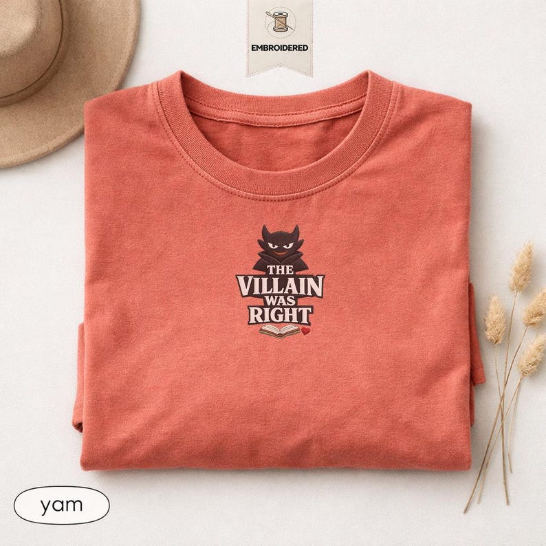 Op de afbeelding: Een terracotta-kleurig T-shirt met ronde hals en een geborduurde afbeelding van een donker, gehoornd personage boven een open boek. De tekst luidt THE VILLAIN WAS RIGHT, vergezeld door een klein rood hartje in het midden op de borst.