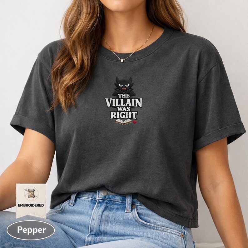 Op de afbeelding: Een antracietgrijs t-shirt met een geborduurde afbeelding van een gestileerd zwart duivelsfiguur boven de tekst "THE VILLAIN WAS RIGHT" en een icoon van een open boek met een klein rood hartje. Het t-shirt heeft korte mouwen en een ronde hals.