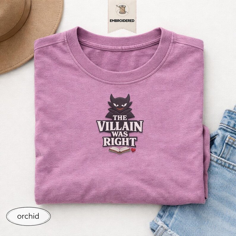 Op de afbeelding: Een orchideekleurig t-shirt met een geborduurd ontwerp van een donker, gehoornd personage boven de tekst "THE VILLAIN WAS RIGHT", een icoon van een open boek en een klein rood hartje. Het ontwerp is gecentreerd op de borst van het kledingstuk.