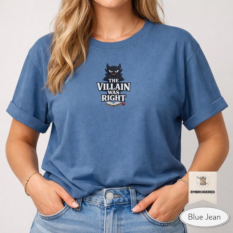 Op de afbeelding: Een blauw t-shirt met korte mouwen en een geborduurde afbeelding op de borst. Het ontwerp bevat een gestileerd zwart wezen boven de witte tekst "THE VILLAIN WAS RIGHT" en een icoon van een open boek met een klein rood hartje.