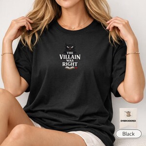 Op de afbeelding: Een zwart t-shirt met korte mouwen en een geborduurde afbeelding op de borst. Het ontwerp bevat een gestileerd donker wezen-icoon boven de witte tekst THE VILLAIN WAS RIGHT, met een klein icoon van een open boek onder de tekst.