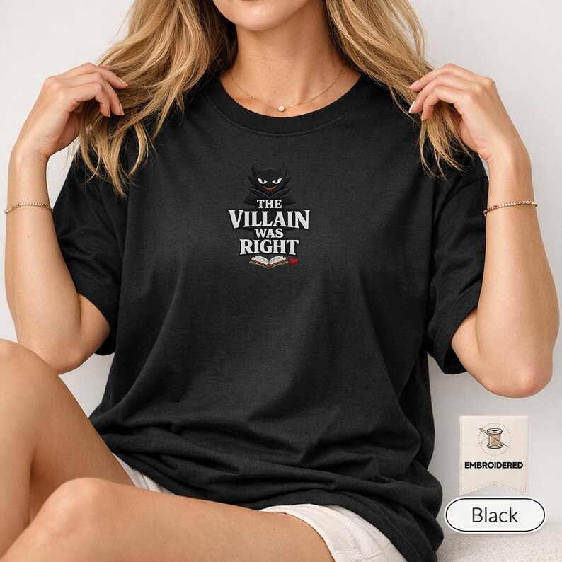 Op de afbeelding: Een zwart t-shirt met korte mouwen en een geborduurde afbeelding op de borst. Het ontwerp bevat een gestileerd donker wezen-icoon boven de witte tekst THE VILLAIN WAS RIGHT, met een klein icoon van een open boek onder de tekst.