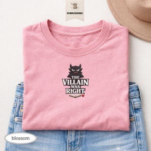 Op de afbeelding: Een lichtroze t-shirt met een geborduurde afbeelding van een zwart gehoornd personage boven de tekst THE VILLAIN WAS RIGHT, een icoon van een open boek en een klein rood hartje. Het shirt is gevouwen over een blauwe spijkerbroek.