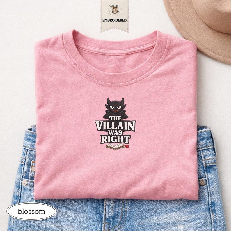 Op de afbeelding: Een lichtroze t-shirt met een geborduurde afbeelding van een zwart gehoornd personage boven de tekst THE VILLAIN WAS RIGHT, een icoon van een open boek en een klein rood hartje. Het shirt is gevouwen over een blauwe spijkerbroek.