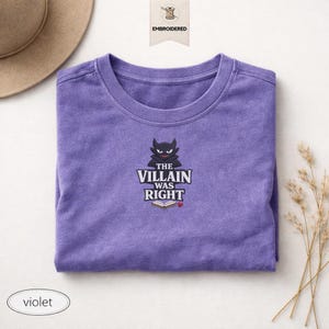 Op de afbeelding: Een violetkleurig t-shirt met ronde hals en een geborduurde afbeelding van een zwart gehoornd personage boven de tekst "THE VILLAIN WAS RIGHT" en een icoon van een open boek met een klein rood hartje. Het t-shirt heeft korte mouwen en een zachte textuur.