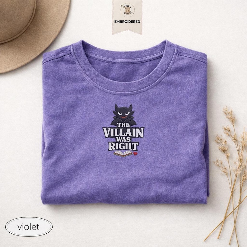 Op de afbeelding: Een violetkleurig t-shirt met ronde hals en een geborduurde afbeelding van een zwart gehoornd personage boven de tekst "THE VILLAIN WAS RIGHT" en een icoon van een open boek met een klein rood hartje. Het t-shirt heeft korte mouwen en een zachte textuur.