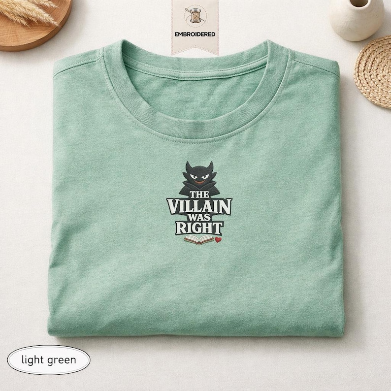 Op de afbeelding: Een lichtgroen t-shirt met ronde hals en een geborduurd ontwerp van een zwart gehoornd personage boven de tekst "THE VILLAIN WAS RIGHT" en een icoon van een open boek met een klein rood hartje. Het ontwerp is gecentreerd op de borst.