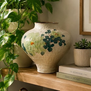 Puede incluir: Un jarrón de cerámica de color crema con una superficie texturizada y un diseño floral pintado en tonos verdes y azules. El jarrón presenta un cuerpo ancho y redondeado que se estrecha hacia la base y un cuello corto y estrecho con borde marrón.