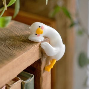 Könnte beinhalten: Eine kleine weiße Gänsefigur mit gelbem Schnabel und orangefarbenen Füßen, die so gestaltet ist, dass sie über die Kante eines Regals gehängt werden kann. Die Gans hat einen glatten, runden Körper, schwarze Augen und rosa Wangenakzente.