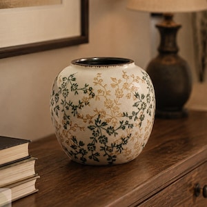 May include: A round ceramic vase with a cream-coloured crackle glaze finish, decorated with a repeating pattern of delicate green and gold floral vines. The vase features a wide, circular opening with a dark, contrasting rim at the top.