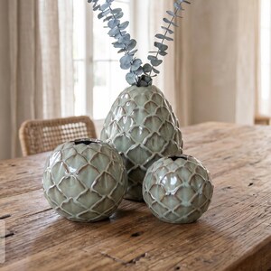 May include: A set of three sage green ceramic vases with a raised, interlocking scale pattern. The tallest vase is oval-shaped and holds two sprigs of dried eucalyptus, while the two smaller vases are spherical in shape with open tops.