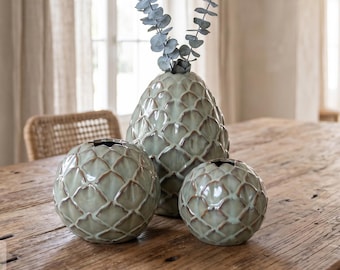 May include: A set of three sage green ceramic vases with a raised, interlocking scale pattern. The tallest vase is oval-shaped and holds two sprigs of dried eucalyptus, while the two smaller vases are spherical in shape with open tops.