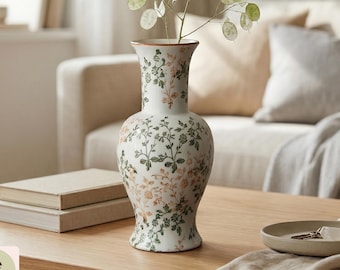 May include: A white ceramic vase with a flared rim and a rounded body, decorated with a repeating pattern of green leaves and orange floral sprigs. A single stem with translucent, circular seed pods is placed inside the vase opening.