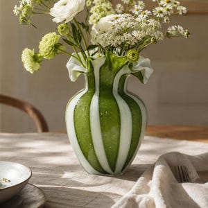 May include: A glass vase featuring vertical stripes of translucent green and opaque white, with a ruffled, flared rim. The vase is filled with a bouquet of white and light green flowers, including ranunculus and small clusters of white blossoms.