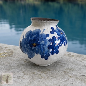 May include: A round ceramic vase with a textured off-white surface featuring hand-painted blue floral motifs. The vase has a wide, slightly flared opening with a dark brown rim and a bulbous body that tapers towards a narrow base.