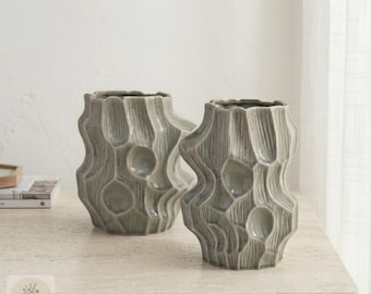 May include: Two sage green ceramic vases with a distinct organic, textured surface featuring vertical ridges and recessed circular indentations. The vases have a glossy finish and a wide, irregular opening at the top, showcasing a sculptural design.