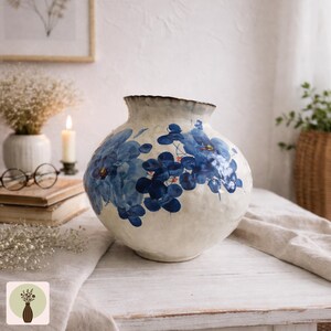 May include: A round, off-white ceramic vase with a wide opening and a textured surface. The vase is decorated with blue floral designs. The rim is a dark brown colour. The vase is on a white wooden table.