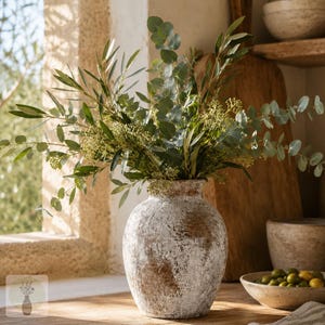 May include: A round, textured ceramic vase with a distressed white and brown finish, filled with a lush arrangement of green eucalyptus branches and olive leaves. The vase has a wide body and a narrow neck, showcasing a weathered, rustic appearance.