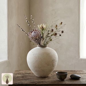 Puede incluir: Un jarr&oacute;n de cer&aacute;mica texturizada de color crema contiene un arreglo floral de flores secas, incluyendo una gran protea de color p&uacute;rpura claro. Un peque&ntilde;o cuenco marr&oacute;n oscuro y una piedra gris oscura lisa est&aacute;n sobre una mesa de madera r&uacute;stica.