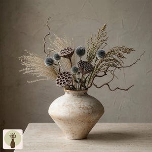 Puede incluir: Un jarr&oacute;n beige texturizado con una forma &uacute;nica, lleno de flores secas, vainas de semillas y hierbas. El arreglo incluye flores espinosas azules parecidas al cardo y elementos delicados y plumosos, creando una est&eacute;tica natural y r&uacute;stica. El jarr&oacute;n est&aacute; sobre una superficie clara.