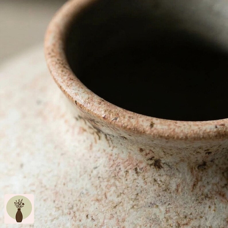 Puede incluir: Primer plano de un jarr&oacute;n de cer&aacute;mica texturizado de color blanquecino con interior oscuro. El borde es marr&oacute;n contrastante con una apariencia moteada. El jarr&oacute;n tiene una est&eacute;tica r&uacute;stica y hecha a mano.