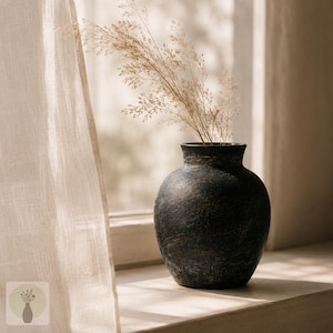 May include: A dark, textured, round-bodied vase with a narrow neck and flared rim. The vase has a matte, weathered black finish with subtle brown undertones and contains a cluster of dried, light beige ornamental grasses with feathery plumes.