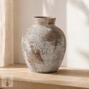May include: A round vase with a wide body and a narrow neck, featuring a distressed finish with layers of white paint over a brown base. The surface has a textured, weathered appearance with visible brush strokes and worn patches throughout the exterior.