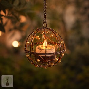 May include: A spherical hanging lantern constructed from dark metal wire, featuring a web-like pattern adorned with small gold-toned beads. A white tea light candle with a warm, glowing flame sits securely in the center, suspended by a metal chain.