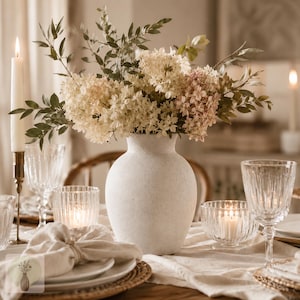 May include: A matte off-white ceramic vase with a rounded body and narrow neck, filled with a bouquet of dried white and pale pink hydrangea flowers and green foliage, positioned as a centerpiece on a table with candles and glassware.