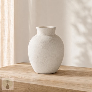 May include: A round, off-white ceramic vase with a textured, matte finish. The vessel features a wide, bulbous body that tapers into a short, narrow neck with a simple, open rim. The surface has a subtle, speckled appearance throughout.