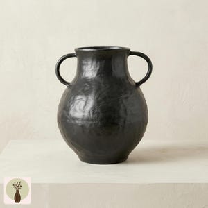 May include: A black ceramic vase with two handles. The vase has a textured surface and a rounded body. The handles are curved and attached to the upper portion of the vase. The vase is on a white surface.