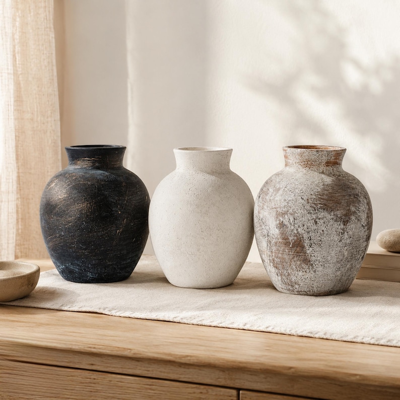 May include: Three rounded vases with narrow necks and textured surfaces. The first vase is dark charcoal with subtle brown highlights, the second is solid off-white, and the third features a distressed white finish over a brown base.