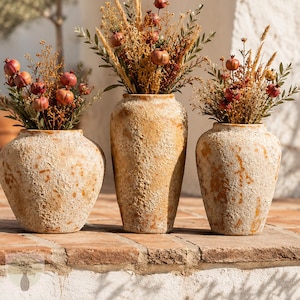 Könnte beinhalten: Drei strukturierte, beige Keramikvasen in unterschiedlichen Formen und Größen, jeweils gefüllt mit getrockneten Blumenarrangements aus kleinen braunen granatapfelartigen Früchten, goldenen Weizenhalmen und grünem Blattwerk, auf einem Sims.