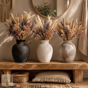 May include: Three rounded vases in black, white, and mottled gray stone textures, each holding a large, dense arrangement of dried flowers, wheat stalks, and pampas grass in shades of purple, beige, and cream, displayed on a wooden bench.