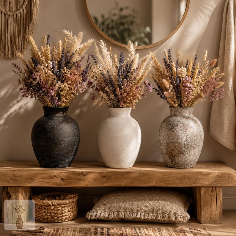 May include: Three rounded vases in black, white, and mottled gray stone textures, each holding a large, dense arrangement of dried flowers, wheat stalks, and pampas grass in shades of purple, beige, and cream, displayed on a wooden bench.