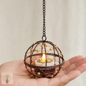 May include: A spherical hanging candle holder crafted from dark brown wire, featuring a delicate web of copper-colored wire accented with small gold beads. A white LED tea light candle sits inside the center, suspended by a dark brown metal chain.