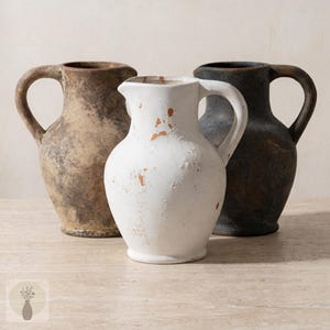 Op de afbeelding: Drie keramische kruiken in verschillende tinten bruin, wit en zwart. Elke kruik heeft een handvat en een tuit. De witte kruik heeft terracotta dat door de verf heen zichtbaar is. De kruiken zijn op een lichtgekleurd oppervlak geplaatst.