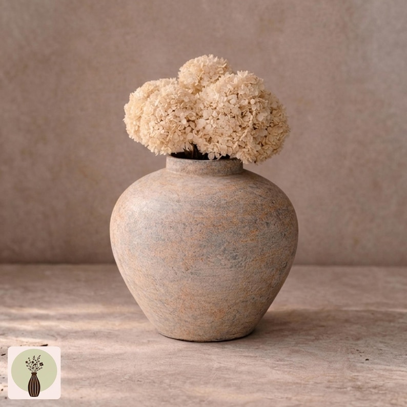 Puede incluir: Un jarr&oacute;n beige texturizado con un cuerpo redondeado y un cuello estrecho, que contiene un racimo de flores secas de color crema. El jarr&oacute;n tiene un aspecto moteado con toques de azul y marr&oacute;n. La est&eacute;tica general es r&uacute;stica y natural.