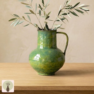 May include: A green and yellow glazed ceramic pitcher with a rounded body, a wide neck, and a single curved handle. The surface features a subtle cross-hatched texture throughout. A leafy olive branch is placed inside the pitcher.
