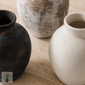 May include: Three round vases with narrow necks and textured surfaces. One vase is matte black with subtle brown undertones, one is off-white with a granular finish, and the third is brown with a distressed white wash finish across the exterior.
