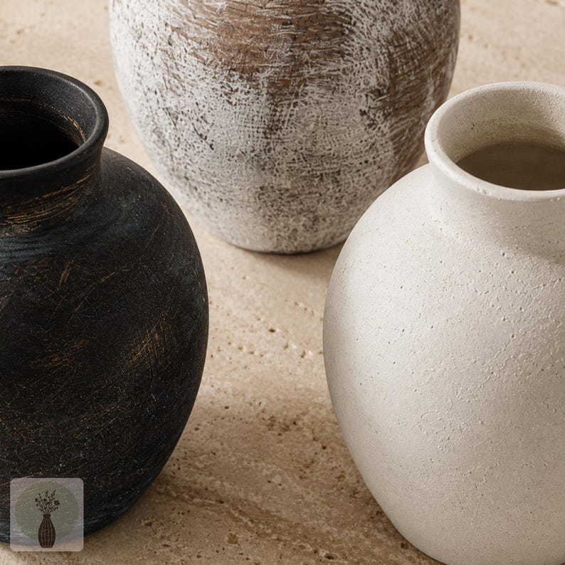 May include: Three round vases with narrow necks and textured surfaces. One vase is matte black with subtle brown undertones, one is off-white with a granular finish, and the third is brown with a distressed white wash finish across the exterior.